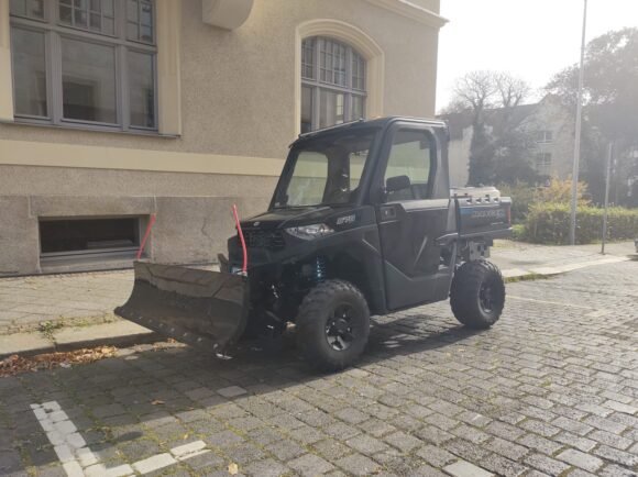 A Polaris Ranger 570 side-by-side UTV parked on a quiet street, fitted with a snow plow.