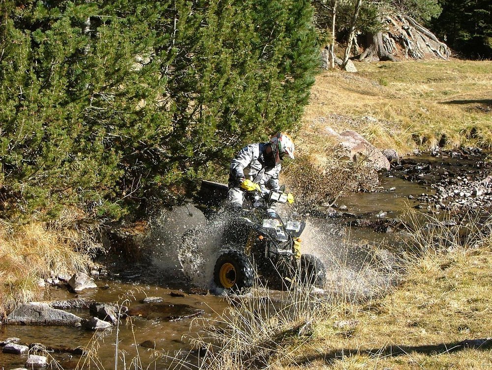 A Polaris Scrambler sport ATV fording a shallow mountain stream with a rider in a white helmet